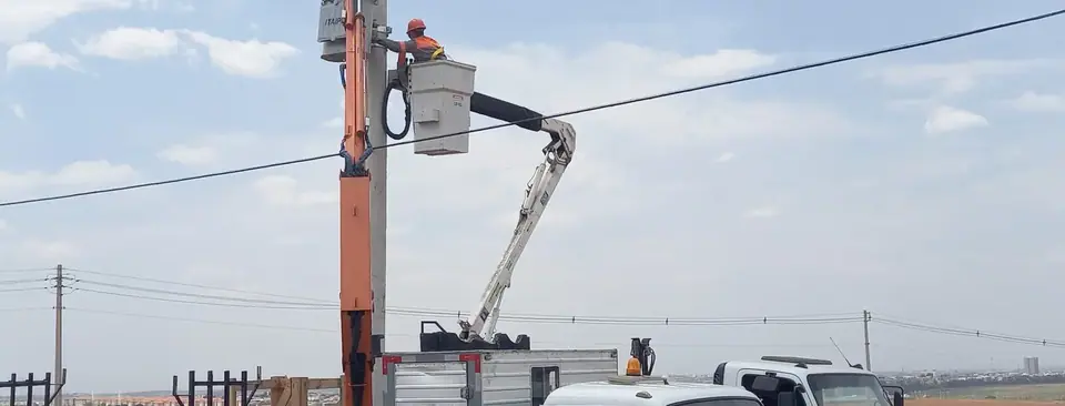 Eletricista da VBE no cesto aéreo trabalhando em rede de distribuição, com os caminhões da empresa